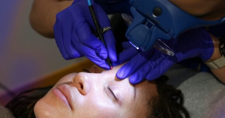 Electrologist in gloves using a fine probe near a client's brow under a magnifying lamp