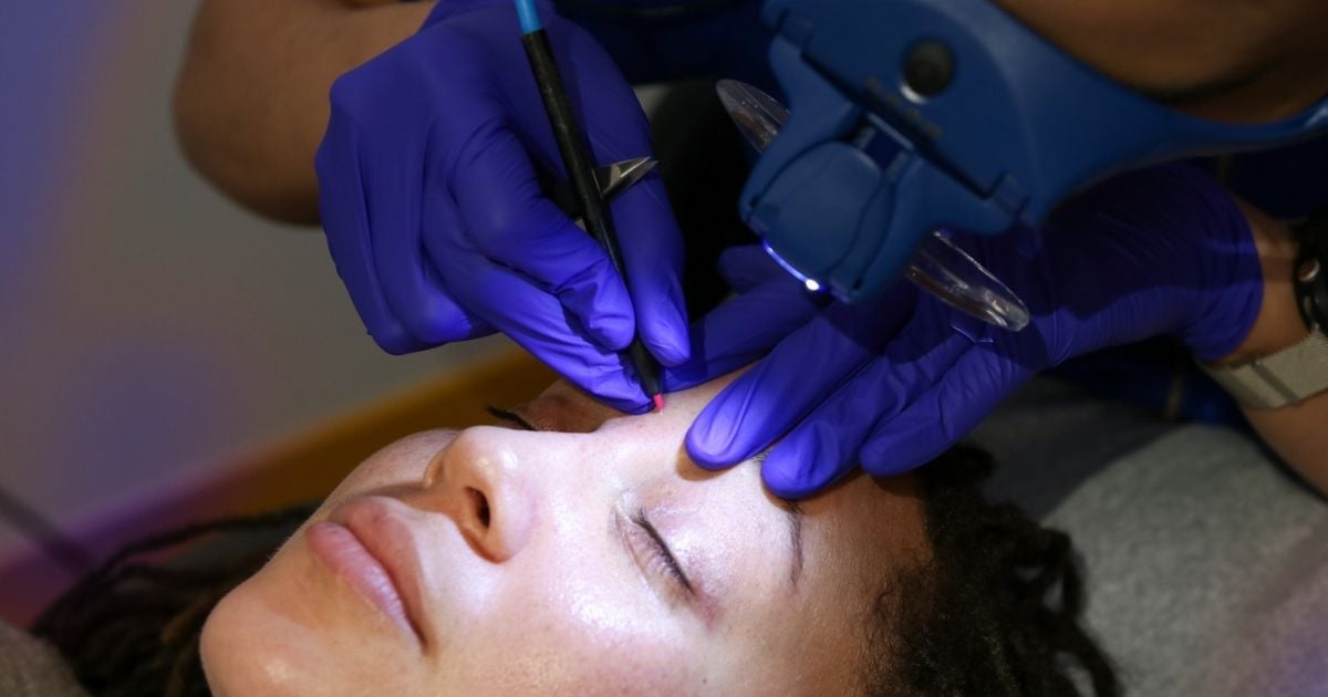 Electrologist in gloves using a fine probe near a client's brow under a magnifying lamp